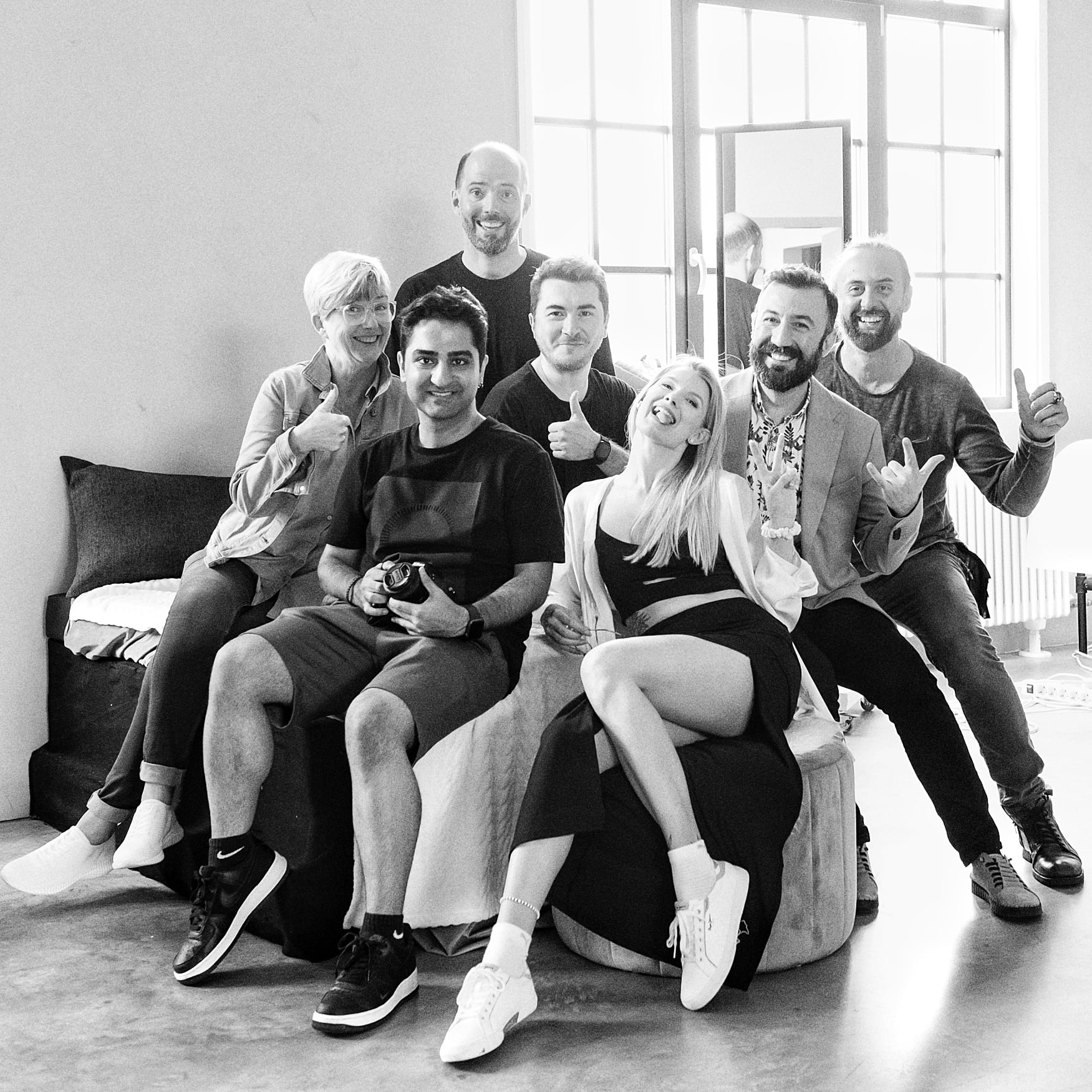 Black-and-white group of seven relaxed participants by window light, playful poses and warm mood; creative fine art nude workshop in Europe.