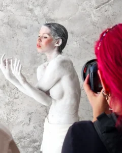 Photographer taking a portrait of a woman covered in white body paint against a textured wall