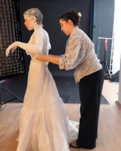 A stylist adjusts the back of a model’s white dress in a studio