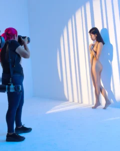 Photographer taking a studio portrait of a nude model against a white wall with striped light and shadow