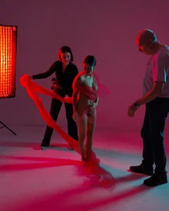 A studio photoshoot with a model being draped in red fabric by two crew members under red lighting