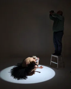 A dancer crouches in a spotlight while a man stands on a stool photographing her in the dark