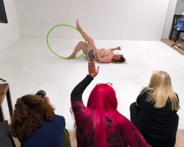 Nude model posing with a hula hoop while three people watch and photograph her in a studio