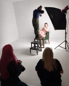 Photography studio shoot with a nude model posing on a chair while a photographer lights the scene and two assistants watch