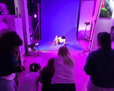 A photographer and onlookers watch a model posing on a purple-lit studio backdrop