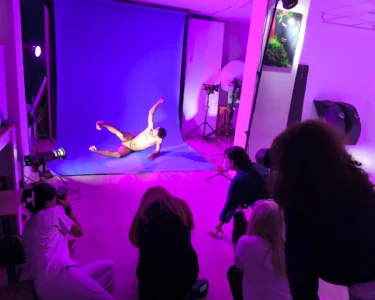 A nude male model posing on a blue backdrop while photographers watch in a studio lit with purple lights
