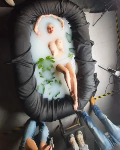 A person lying in a milky bath with arms raised, surrounded by black padding and several people standing around them