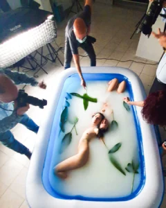 Woman posing in a milk bath with green leaves while photographers take pictures around her