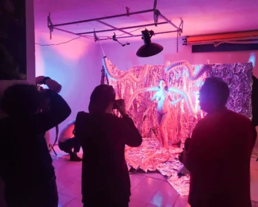 Photographers shooting a nude model posed in front of a pink foil backdrop under studio lights