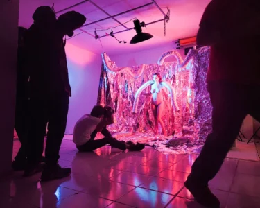 Photo shoot in a pink-lit studio with a model posing in front of a foil backdrop while photographers work around her