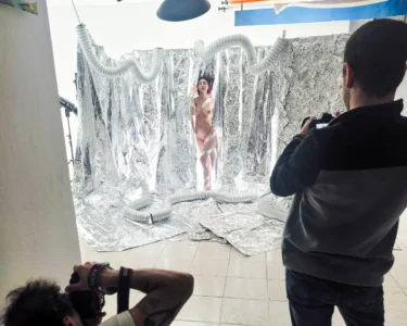 A nude model posing in a silver foil photo studio while two photographers take pictures