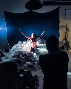 A nude model kneels in a cloud-like set with arms raised during a studio photo shoot