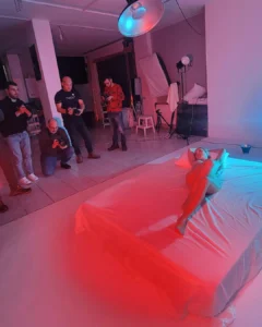 A nude model posing on a bed during a studio photo shoot while photographers watch and take pictures