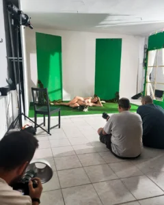 Photo shoot in a studio with a nude model posing on a green backdrop while photographers take pictures