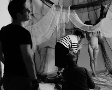 Black-and-white photo shoot with a nude model and crew arranging draped fabric and netting