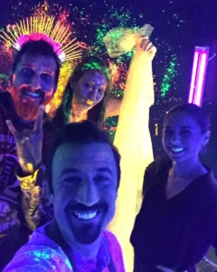 Four people posing under blacklight at a neon party, with glowing face paint and accessories