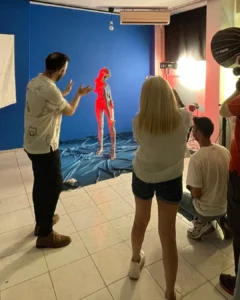 Photoshoot in progress with a model posing against a blue backdrop while crew members direct and light the scene